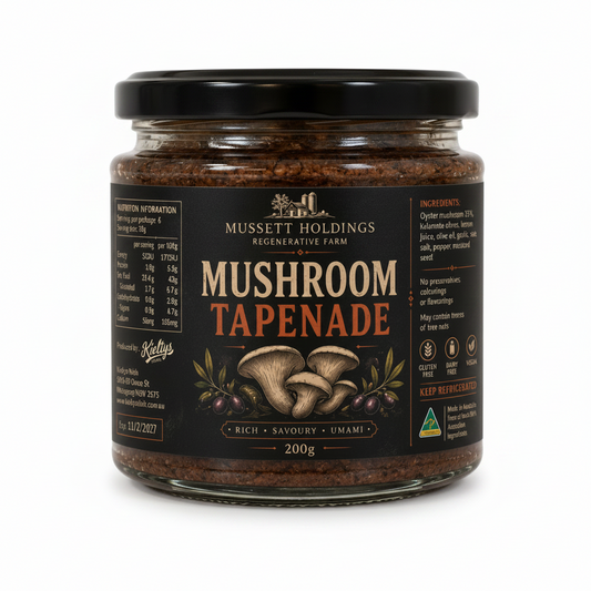 A jar of Mussett Holdings Mushroom Tapenade, 200g, on a light countertop with a blurred kitchen background. The product is in a clear glass jar with a black lid and a black label featuring brown and orange text and mushroom illustrations.